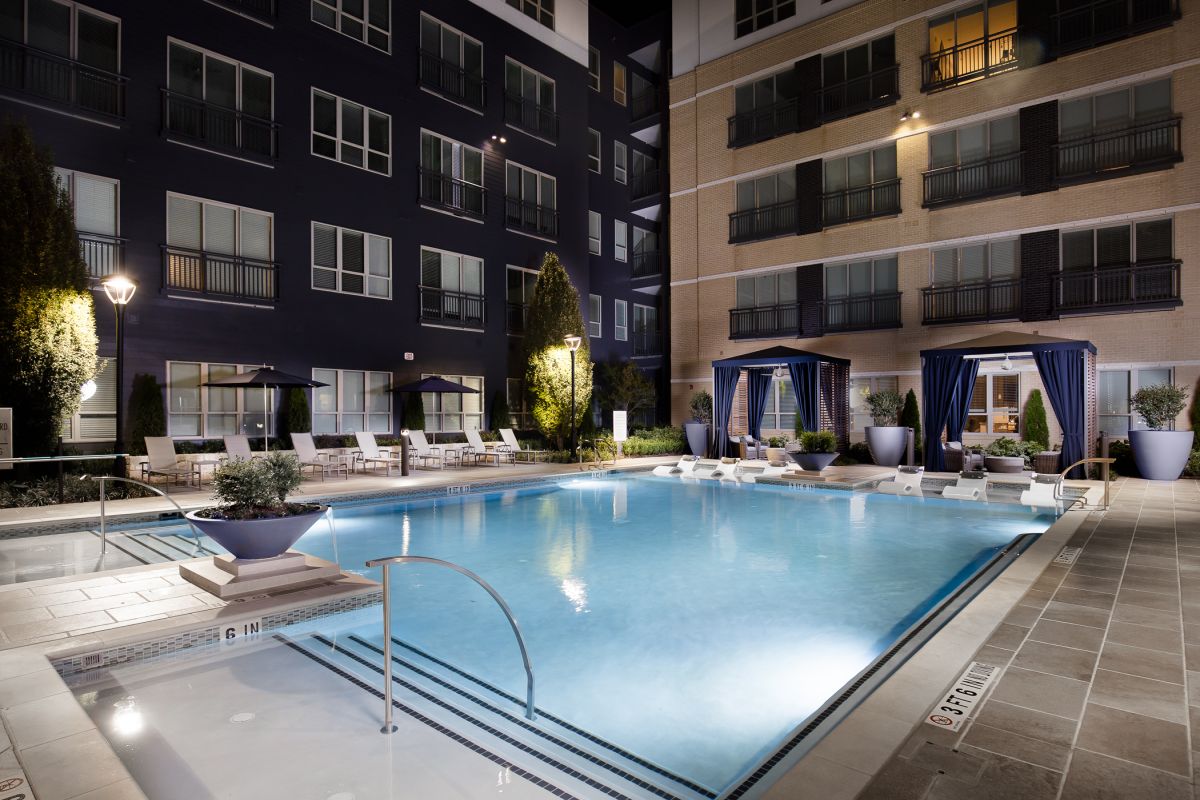 Night view of a modern apartment pool area with lounge chairs, cabanas, and potted plants.