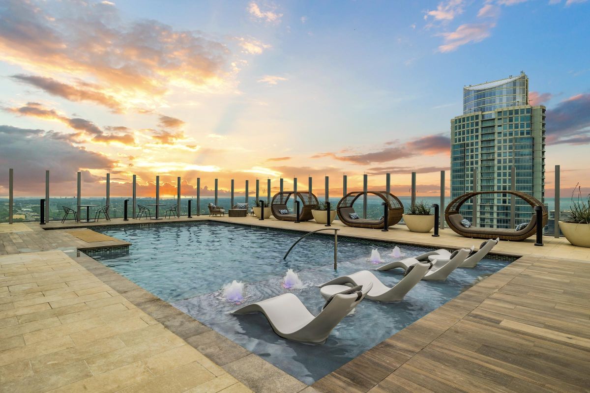 Rooftop pool with lounge chairs and circular seating at sunset, overlooking a cityscape with a tall building.