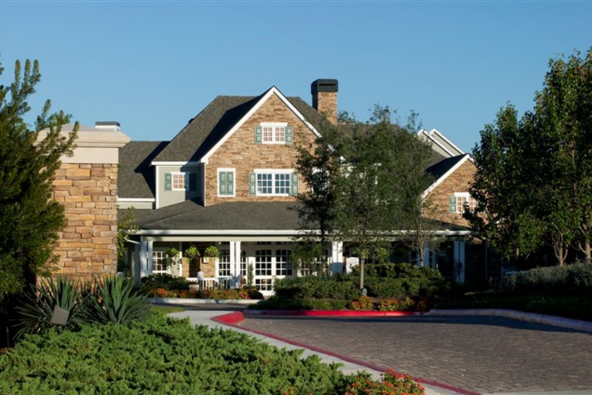 Two-story brick house with a front porch, surrounded by trees and landscaping, under a clear blue sky.
