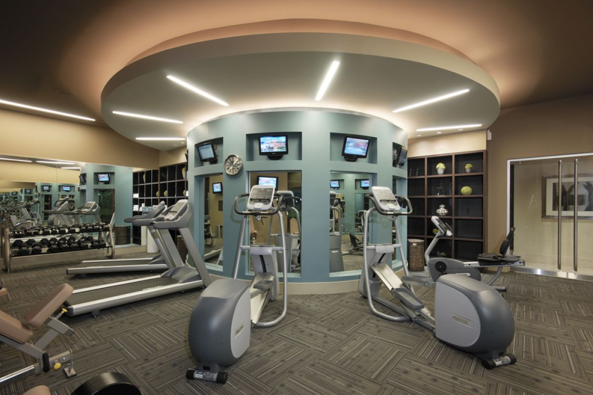 Modern Ashton gym with treadmills, exercise bikes, TVs on a curved wall, mirrors, and shelves with equipment.