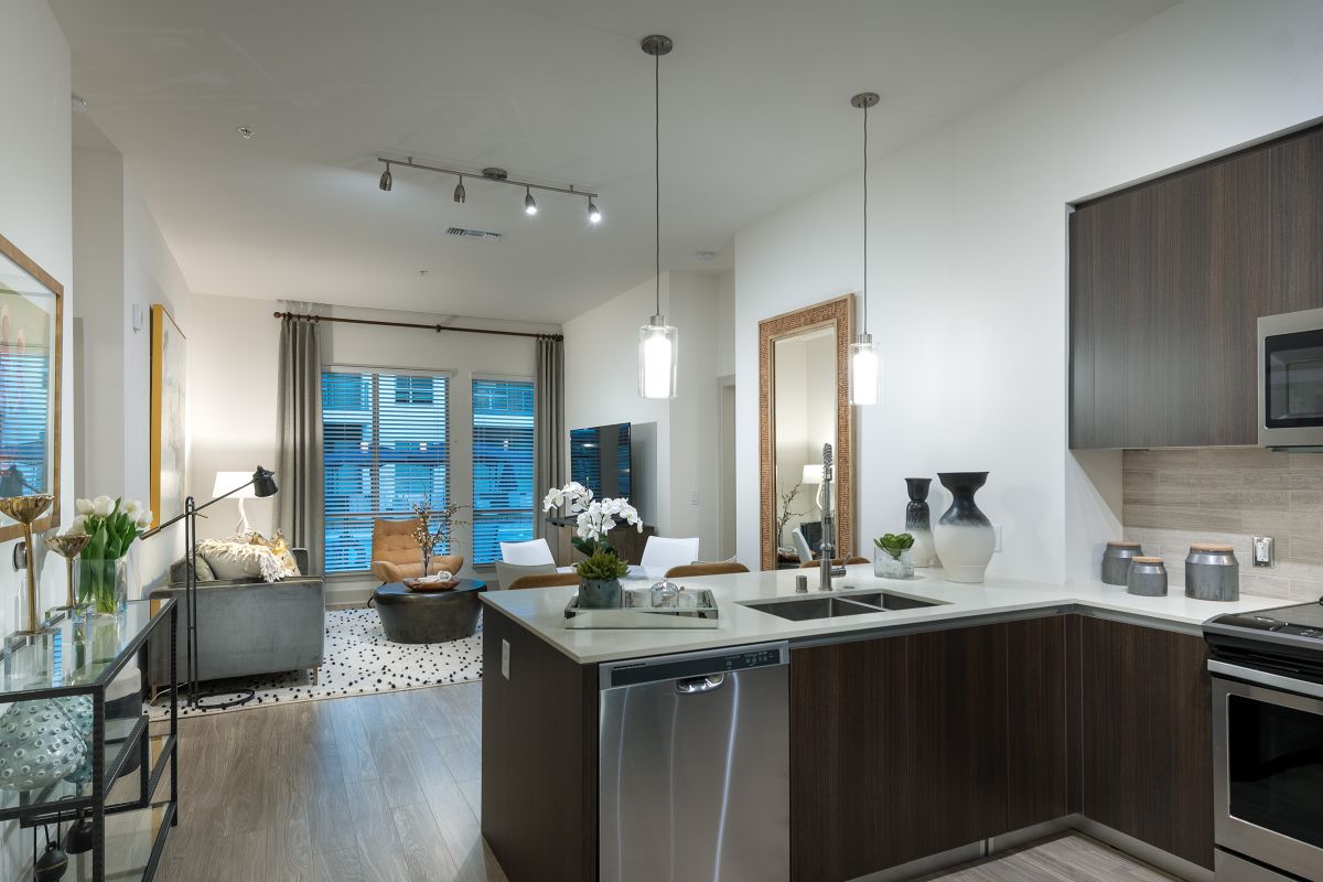 Modern kitchen and living room with dark cabinets, pendant lights, and large windows with blinds.