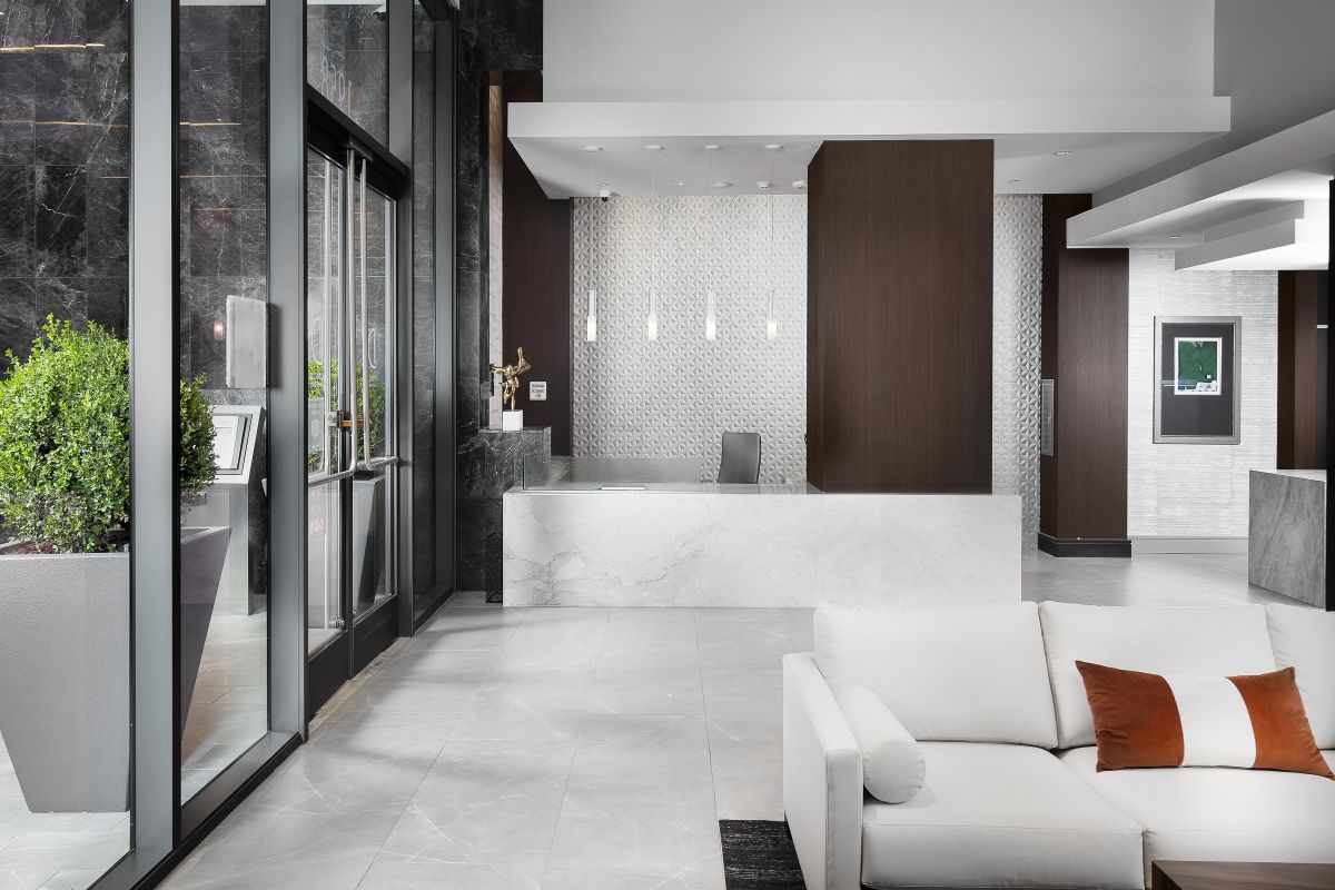 Modern lobby with a white sofa, marble reception desk, and large windows. Contemporary decor with plants and wall art.