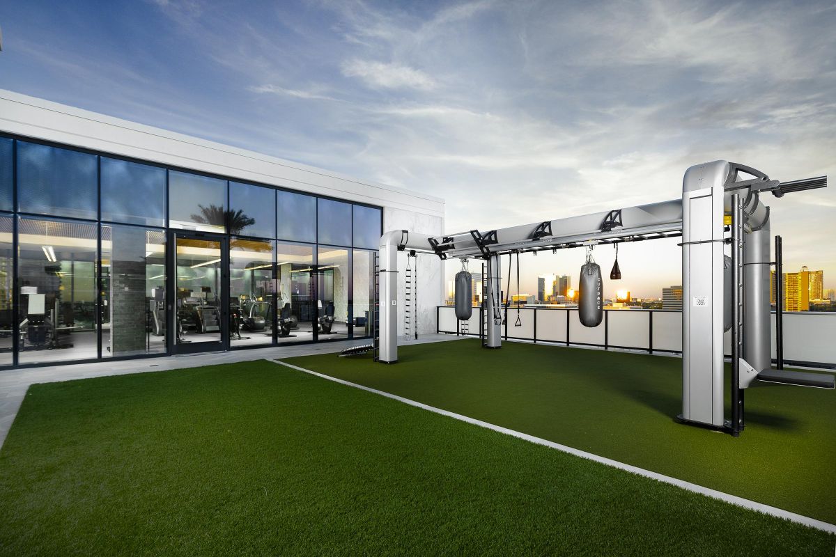 Rooftop gym with punching bags, green turf, and city skyline view at sunset. Glass-walled interior gym visible.