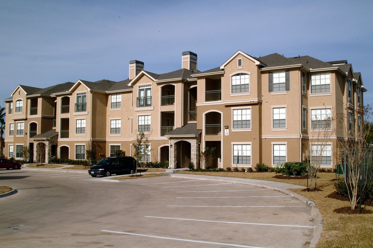 A modern, beige-colored apartment building with multiple windows and a mostly empty parking lot in front.