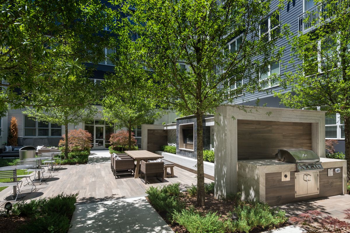 Modern outdoor patio with a grill, dining tables, greenery, and tall trees beside an apartment building.