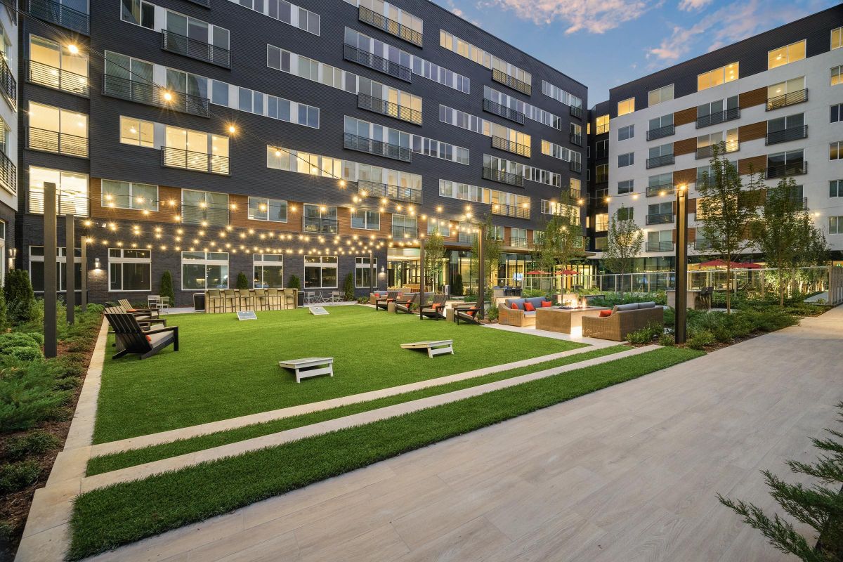 Modern apartment courtyard with string lights, green lawn, cornhole boards, and seating areas at dusk.