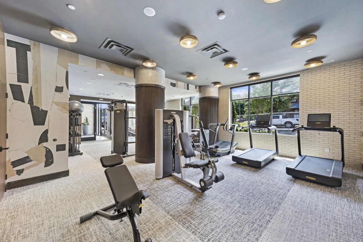 Modern gym with exercise machines, including treadmills, elliptical, and weights, featuring large windows and bright lighting.
