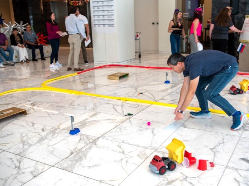 A man adjusts a toy truck on a marble floor track as people watch and talk in the background.