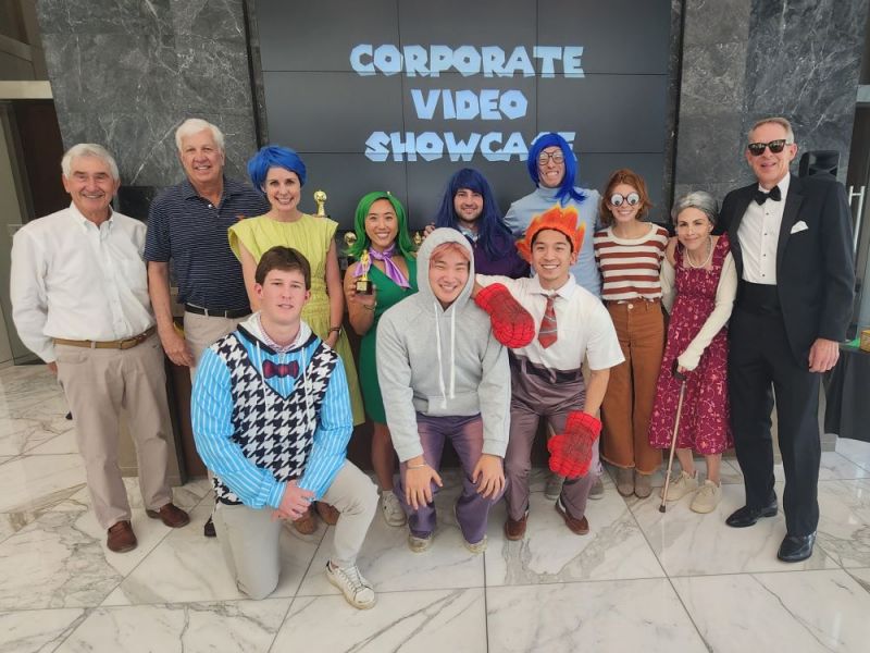 A group of people in costumes pose together in front of a "Corporate Video Showcase" sign.
