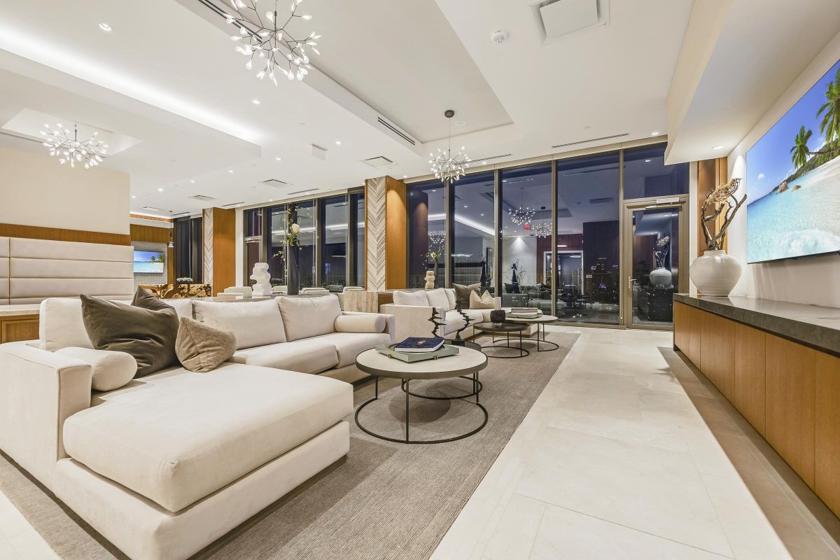 Luxury living room with beige sofas, modern chandeliers, large windows, and a beach scene on a wall-mounted TV.