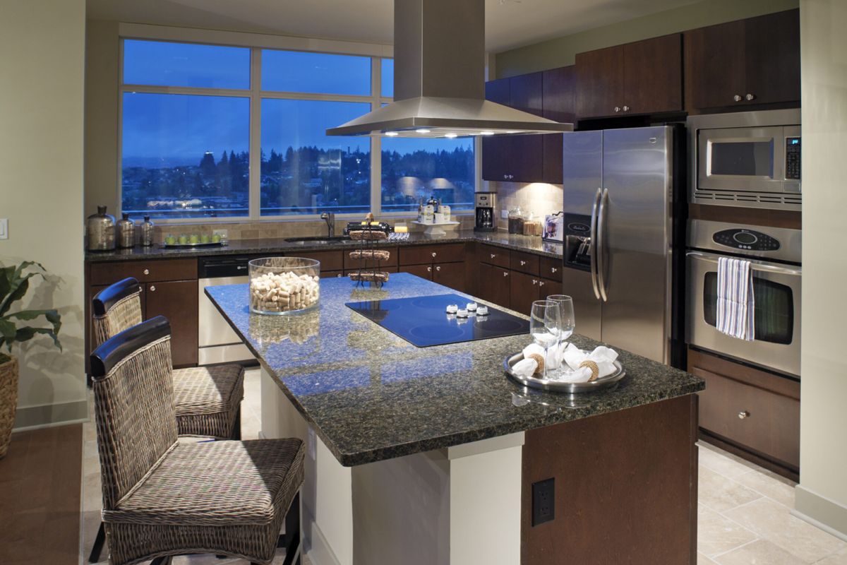 Modern kitchen with granite island, wicker chairs, stainless steel appliances, and large evening windows.
