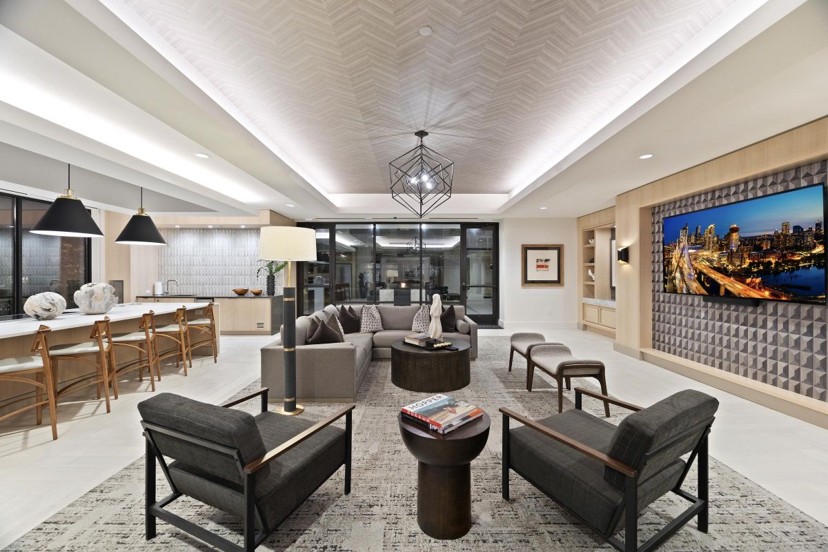 Modern open-concept living room with kitchen, gray sofas, armchairs, and wall-mounted TV displaying cityscape.