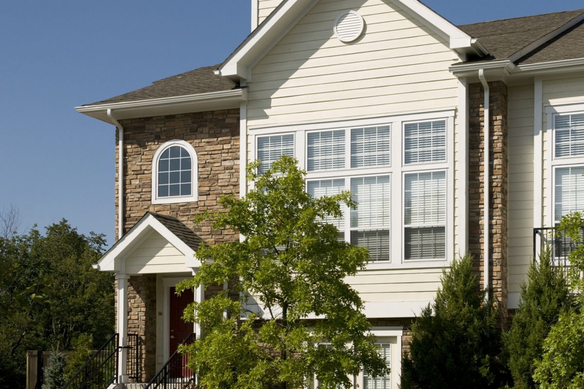 Two-story suburban house with tan siding, stone accents, large windows, and a tree in front.
