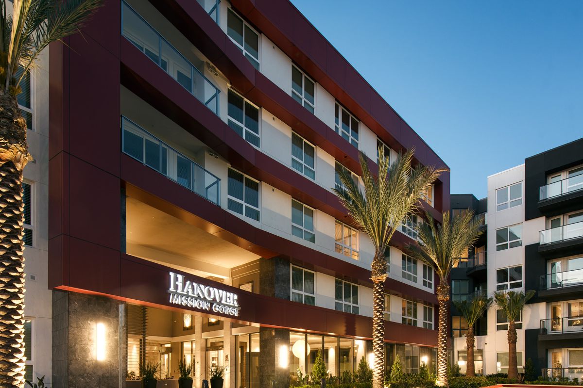 Modern apartment building with palm trees at entrance, lit up during dusk, sign reads "Hanover Mission Gorge.