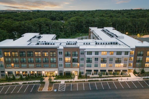 Aerial view of a modern apartment building with surrounding parking and forest in the background at sunset.