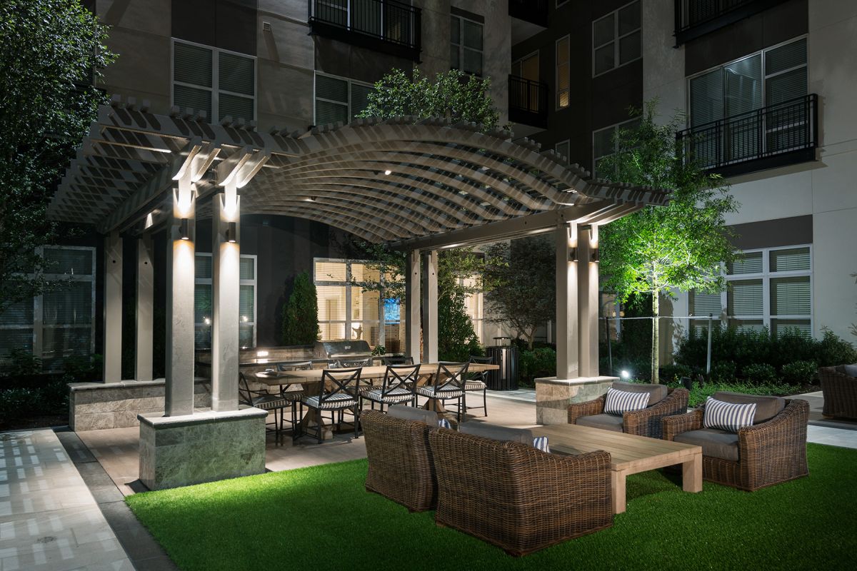 Modern patio at night with wicker seating, dining table under a pergola, and soft lighting on artificial grass.