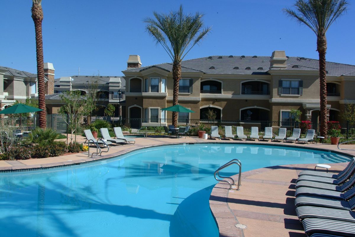 Curved outdoor swimming pool with lounge chairs, palm trees, and apartment buildings in the background.