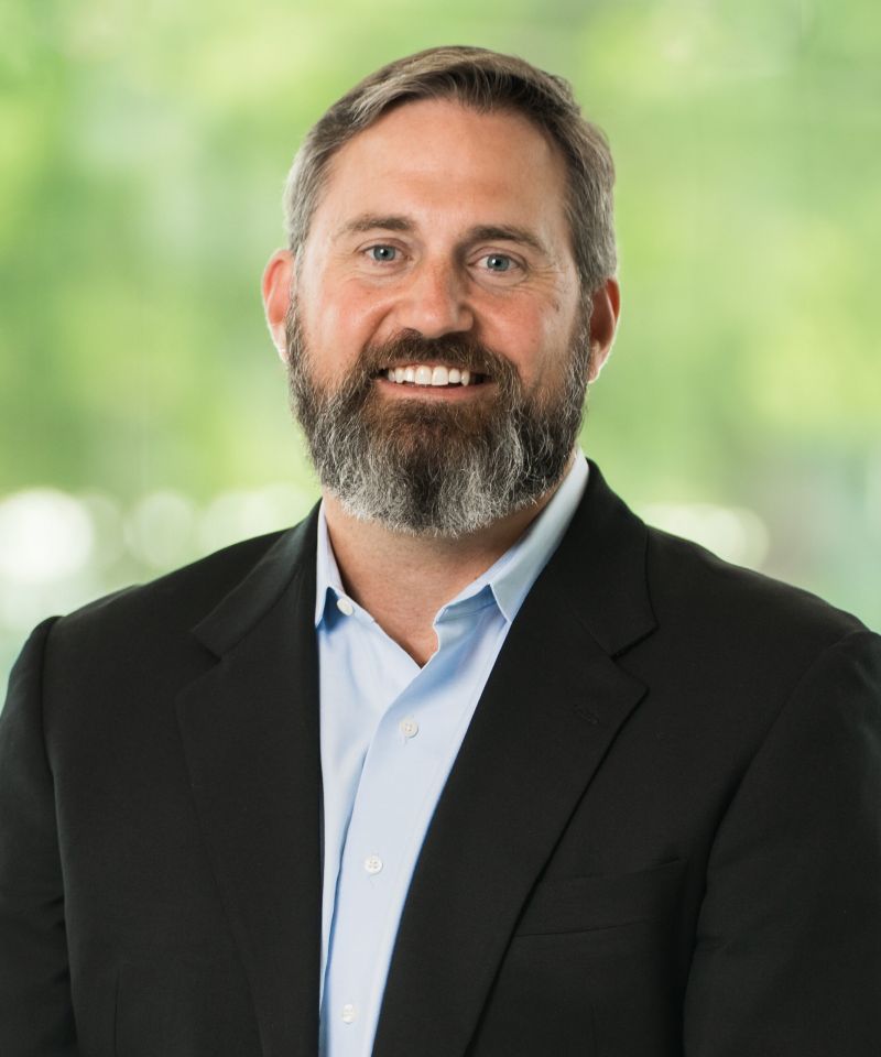Smiling man with short hair and a beard, wearing a black blazer and light blue shirt, in front of a green background.