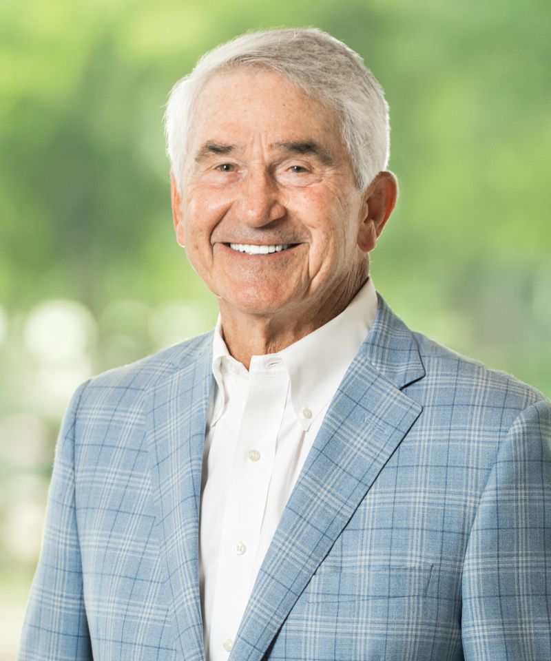 Smiling older man in a light blue plaid suit jacket and white shirt, with a blurred green background.