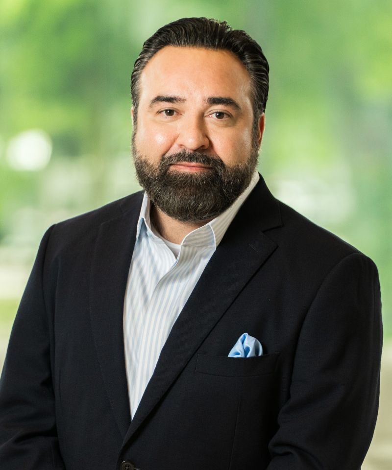 Man with a beard in a dark suit and striped shirt, standing in front of a blurred green background.
