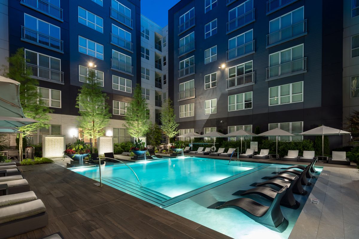 Modern apartment courtyard with a lit swimming pool, lounge chairs, umbrellas, and surrounding greenery at night.