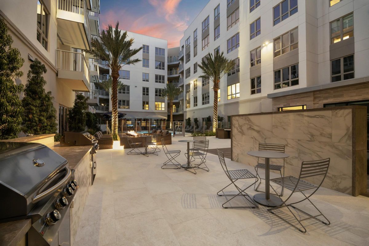 Modern outdoor courtyard with seating, grills, palm trees, and lit fire pits at dusk.