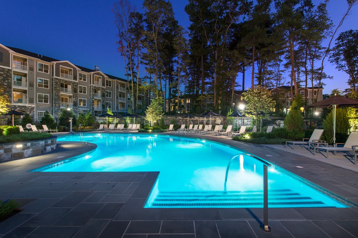 Illuminated outdoor swimming pool at dusk, surrounded by lounge chairs, umbrellas, and apartment buildings.