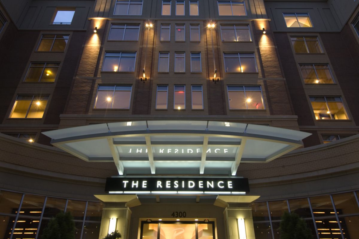 Modern apartment building entrance at night, lit sign reading "THE RESIDENCE" above glass doors.