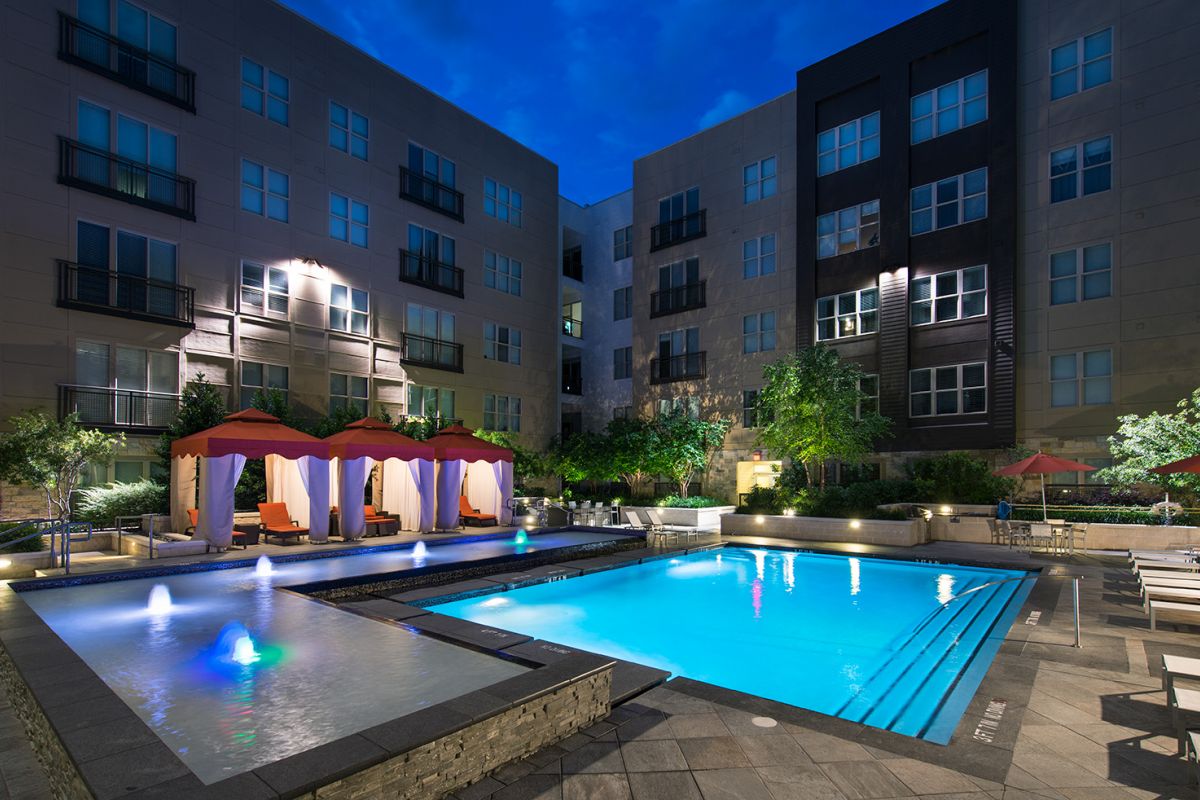 Modern apartment courtyard with illuminated swimming pool, lounge chairs, and cabanas at night.