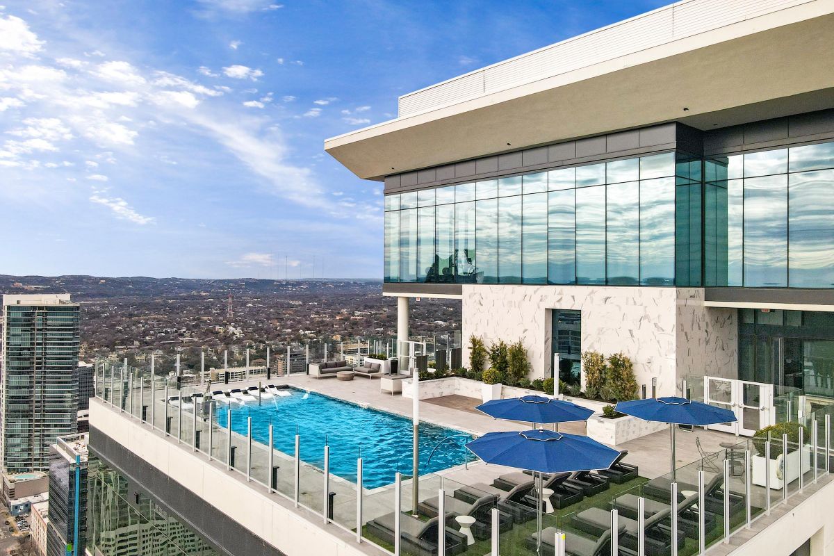 Rooftop pool with lounge chairs and blue umbrellas overlooking Republic Square and the Hanover city skyline under a clear sky.