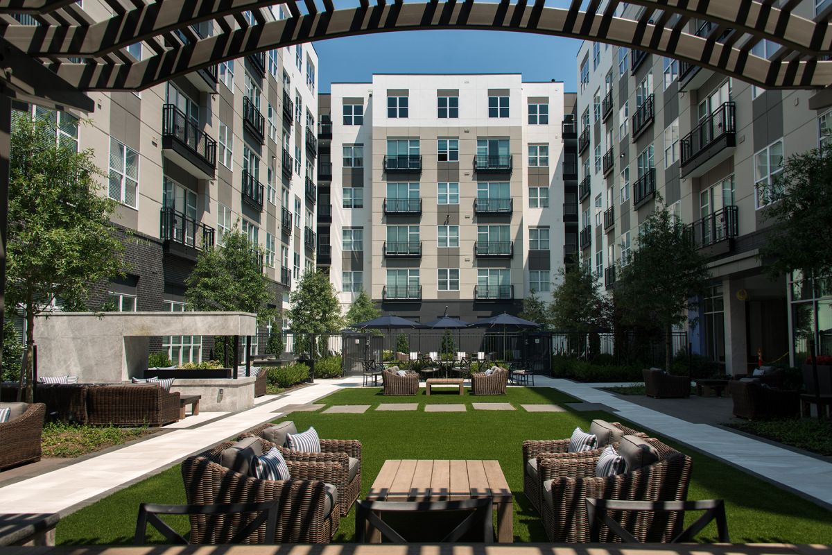 Modern apartment courtyard with outdoor seating, green lawn, trees, and patio umbrellas under a striped pergola.