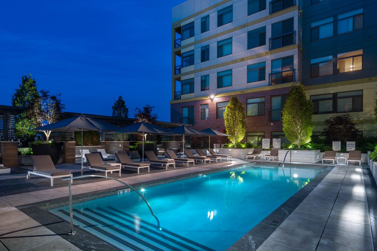 Outdoor swimming pool at night with lounge chairs, umbrellas, and lights beside a modern apartment building.