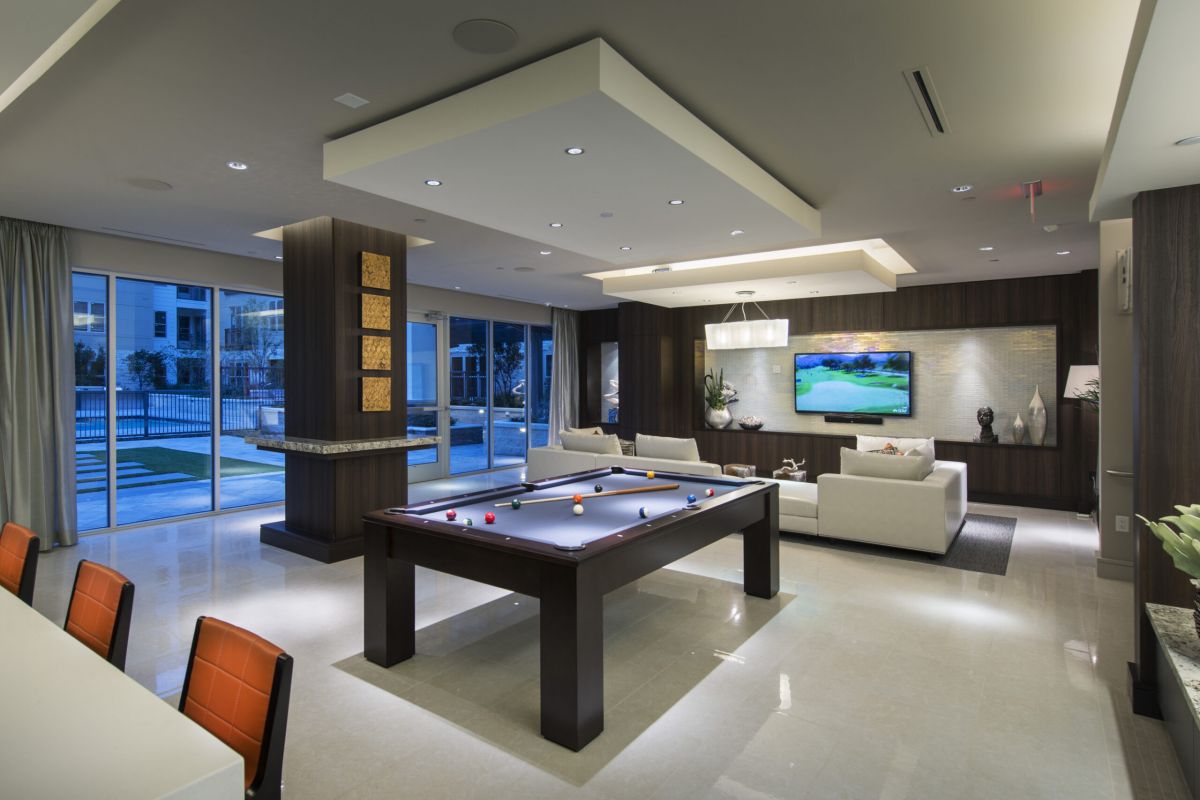 Modern lounge with a pool table, white sofas, TV, and large windows overlooking an outdoor pool area.