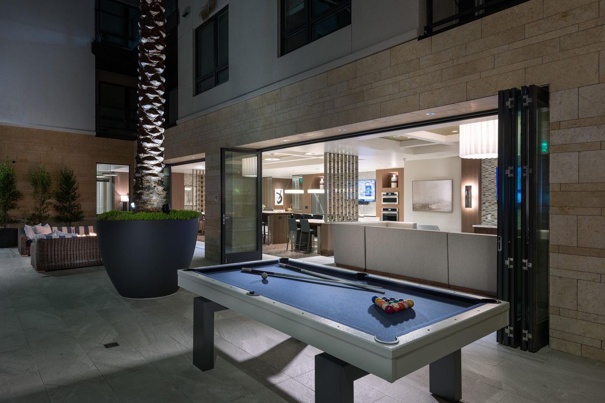 Outdoor pool table beside a large planter, with indoor lounge area visible through open glass doors at night.