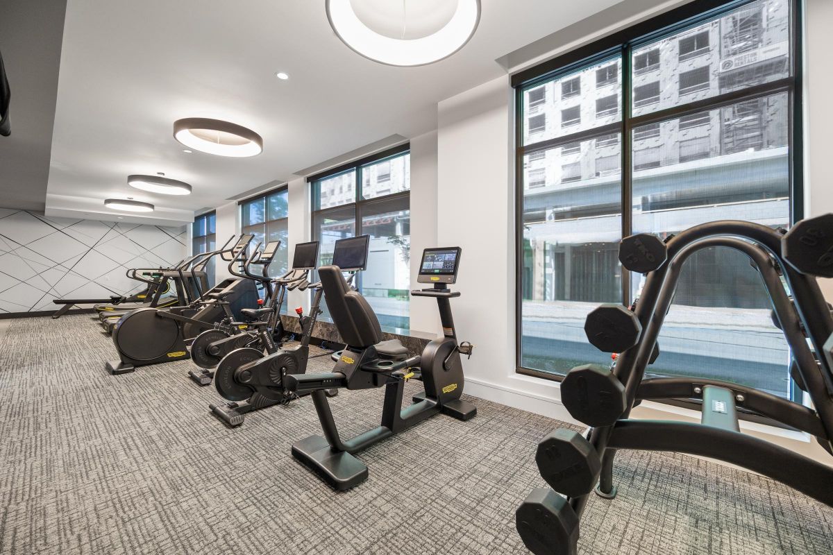 Modern gym with stationary bikes, elliptical machines, and weights near large windows overlooking a cityscape.