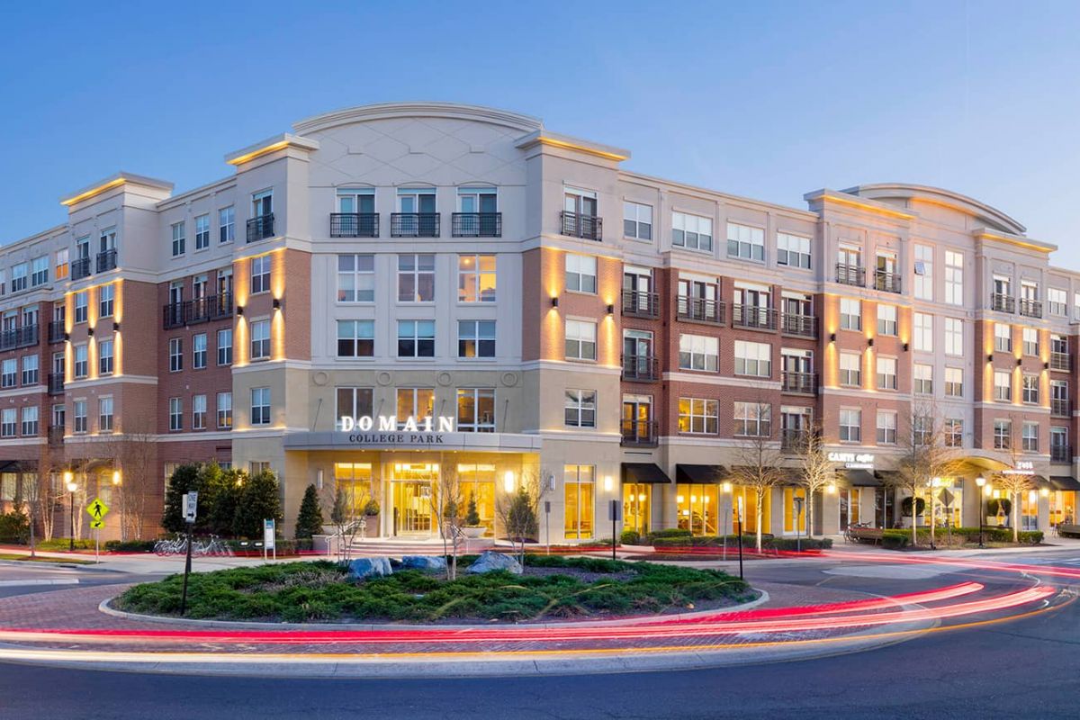Modern, multi-story apartment building with retail shops at dusk, illuminated by streetlights and car light trails.