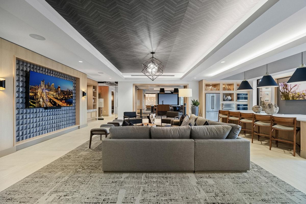Modern living room with a gray sofa, large TV, open kitchen, barstools, and patterned ceiling.