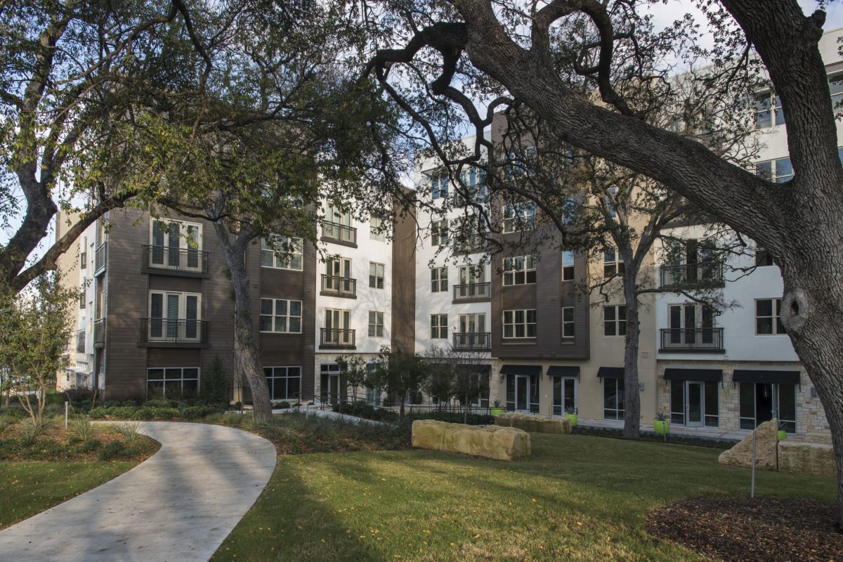 A curved path leads through a grassy area with large trees toward modern apartment buildings.