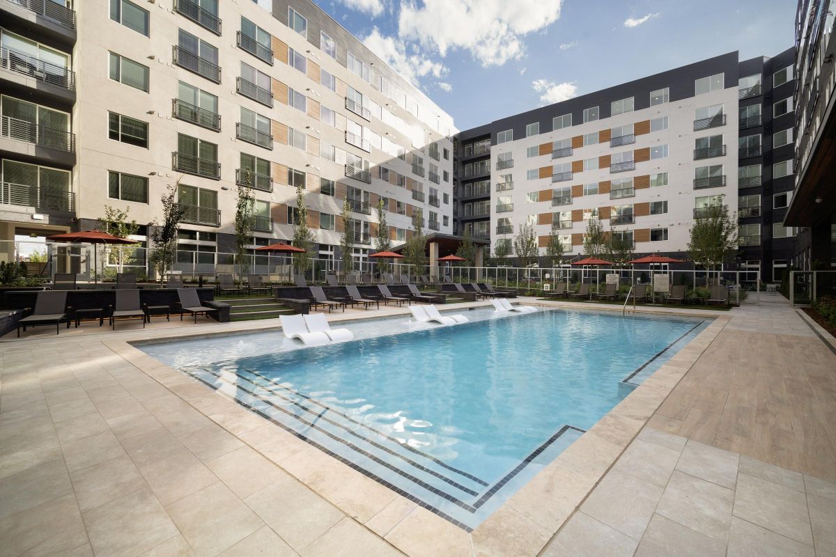 Modern apartment complex featuring a swimming pool, sun loungers, and seating with umbrellas on a sunny day.