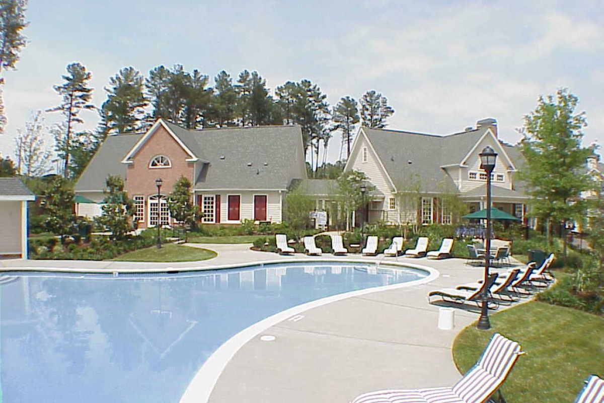 Curved outdoor pool with lounge chairs, trees, and large houses in the background under a clear sky.