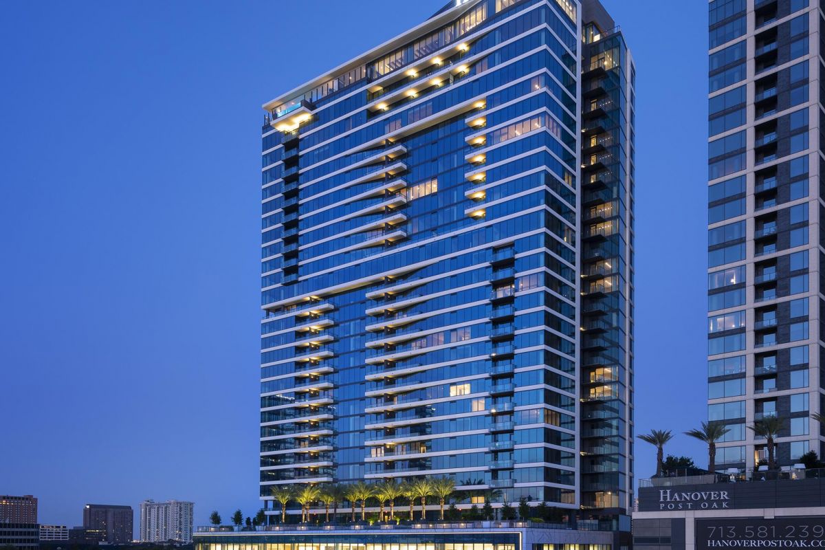 Tall, illuminated modern glass building at dusk with "Hanover" signage, surrounded by other buildings and trees.