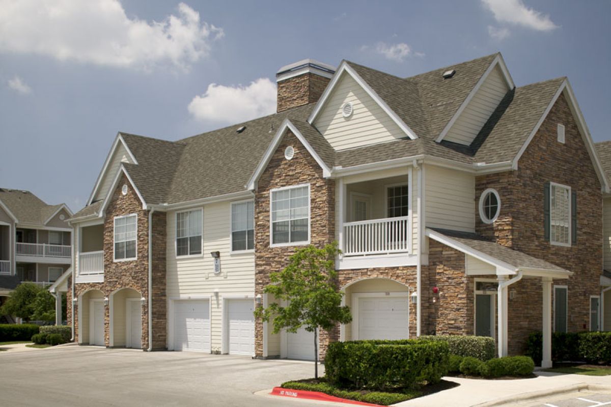 Two-story suburban townhouse building with stone facade, garages, and trimmed bushes under a partly cloudy sky.