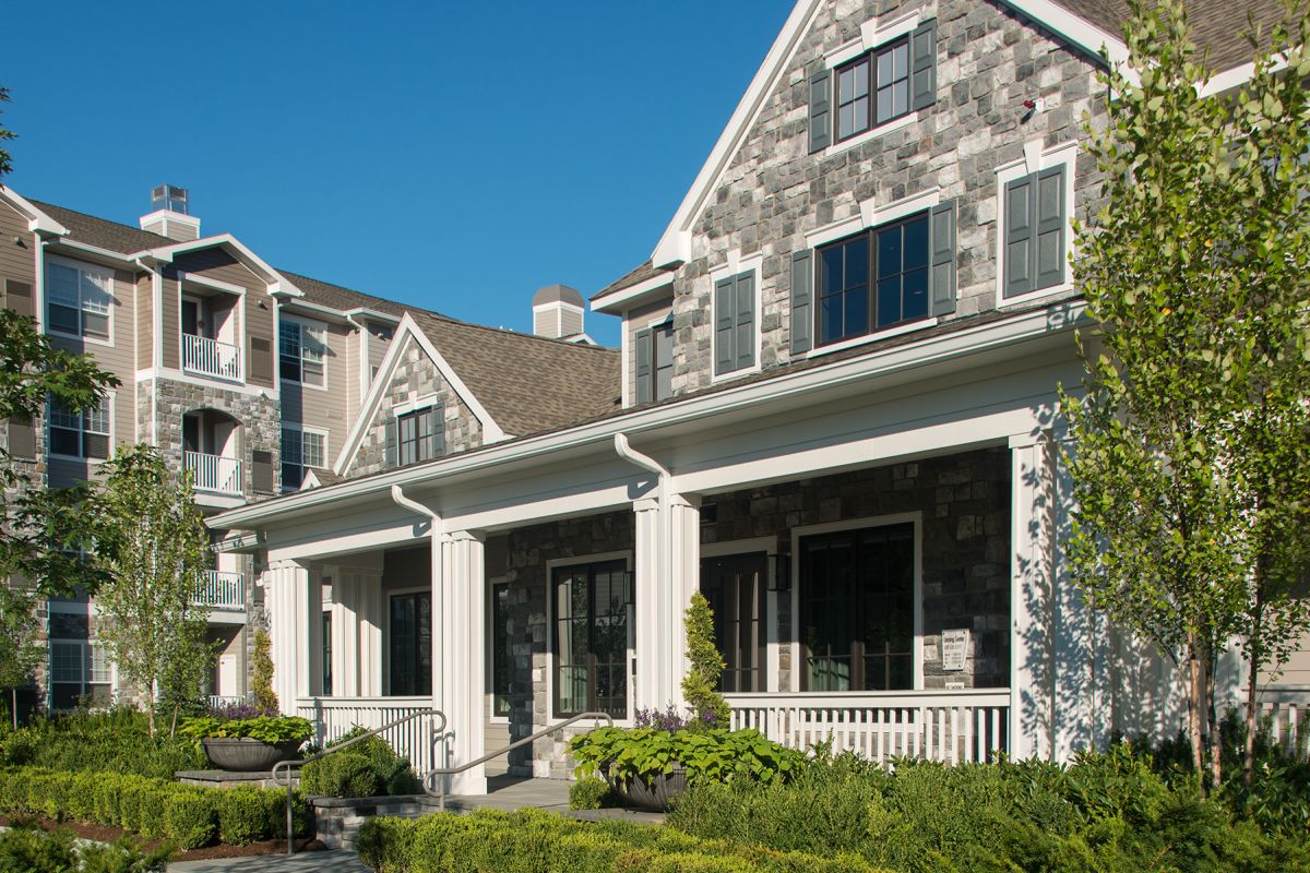 A modern apartment building with stone and siding exterior, columns, and landscaped greenery under a clear sky.