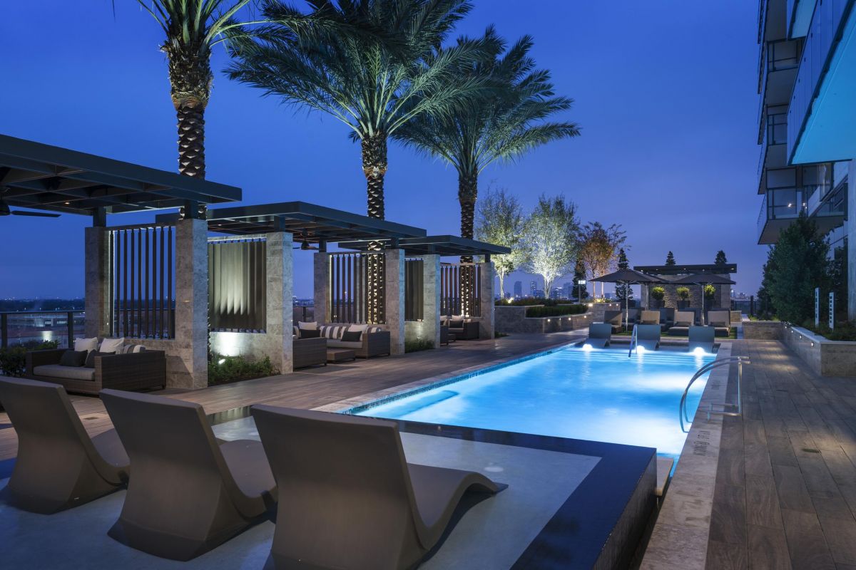 Rooftop pool area at night with lounge chairs, palm trees, and city view.