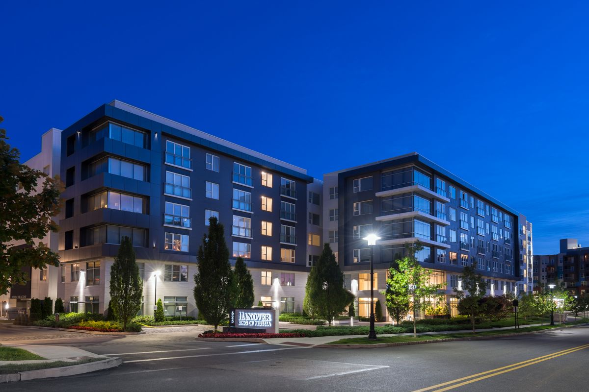 Modern apartment building at dusk, with illuminated windows and a landscaped entrance along a quiet street.