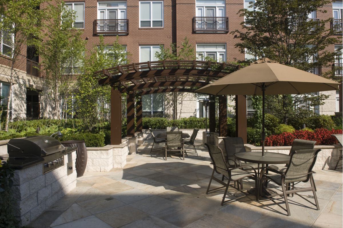 Outdoor patio with tables, chairs, a grill, pergola, and umbrella at Charles River Landing near modern apartments and greenery.