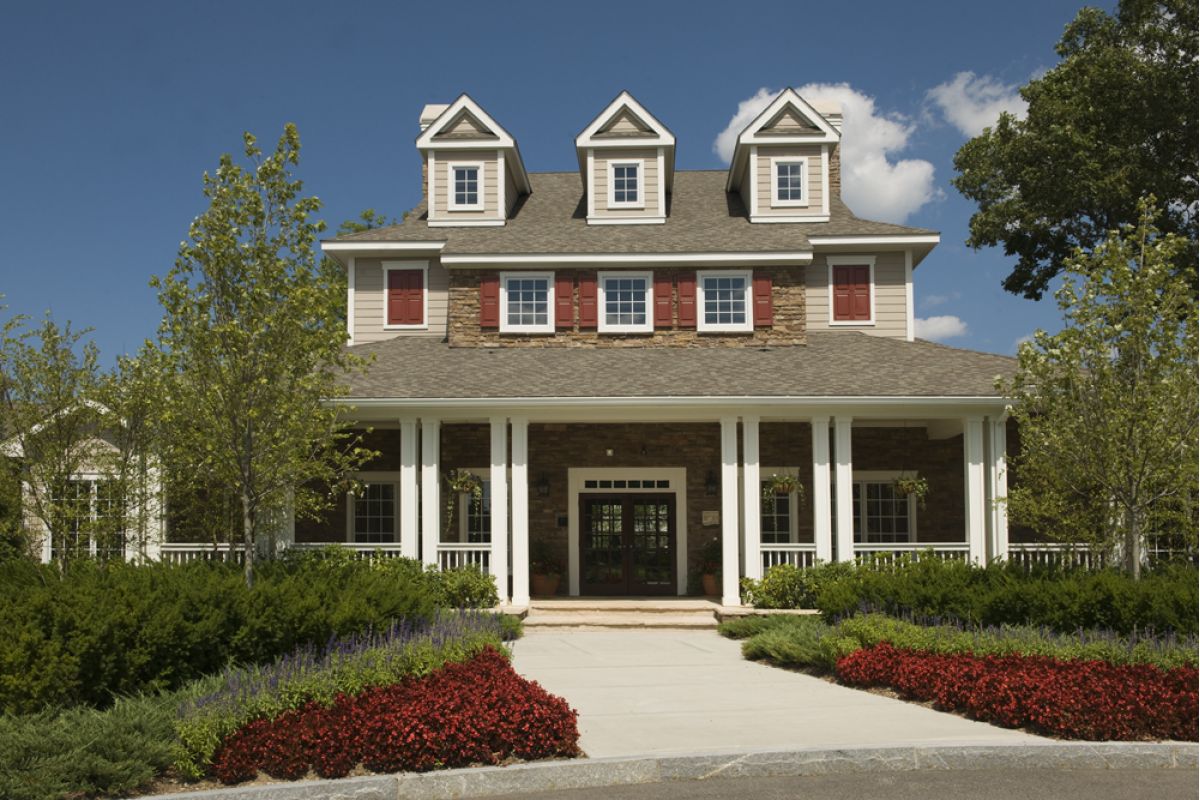 Large house with three dormer windows, red shutters, and a columned porch, surrounded by colorful gardens.