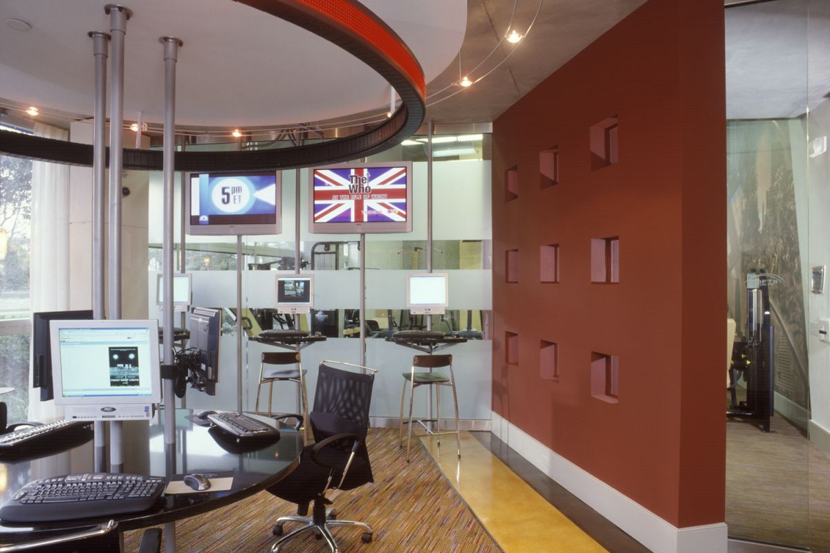 Modern office with computers, glass walls, red accent wall, and TVs displaying news and a UK flag.