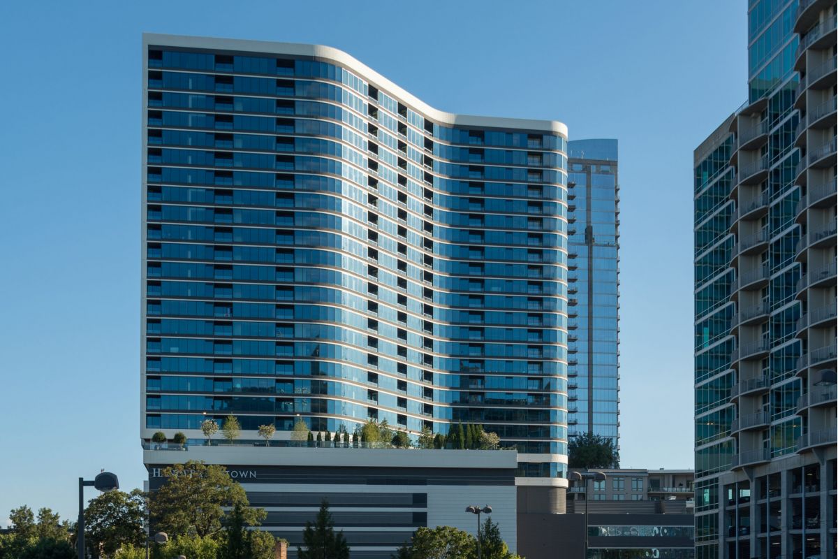 Curved modern high-rise building with reflective glass windows, surrounded by trees and other tall buildings.