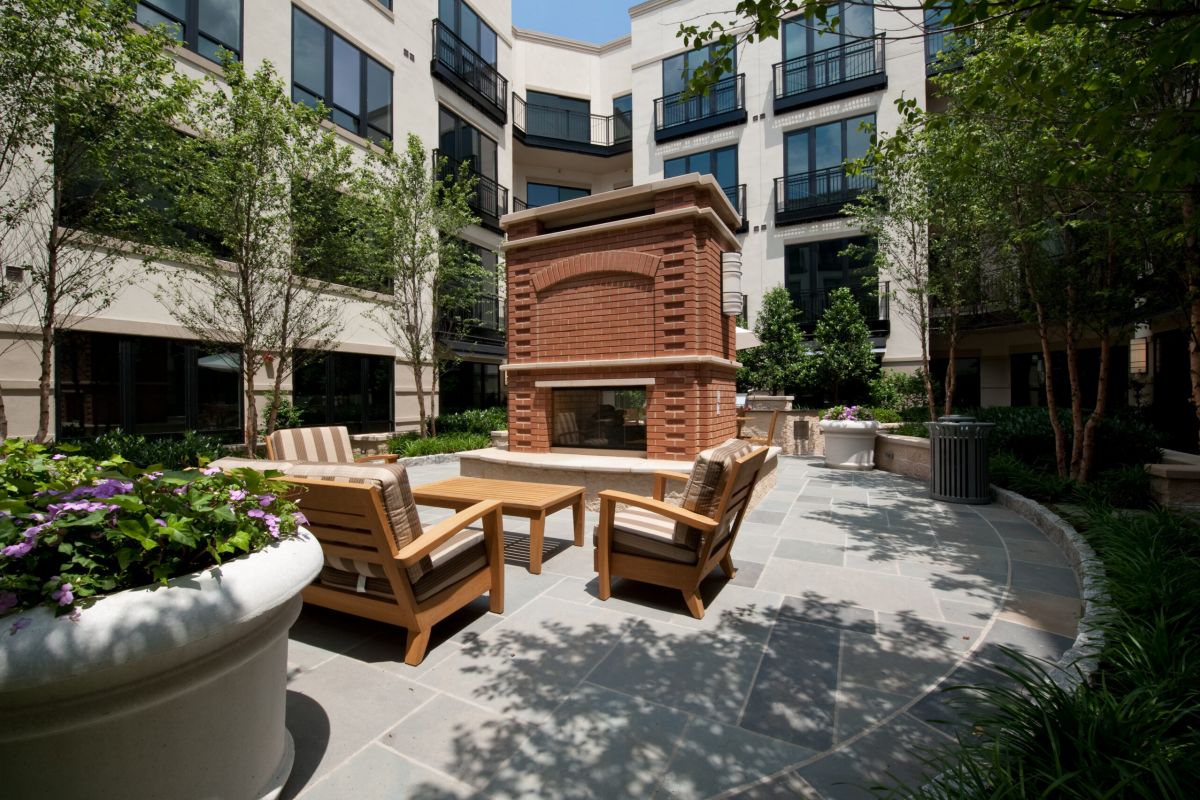 Outdoor courtyard with a brick fireplace, wooden seating, potted flowers, and surrounding apartment buildings.